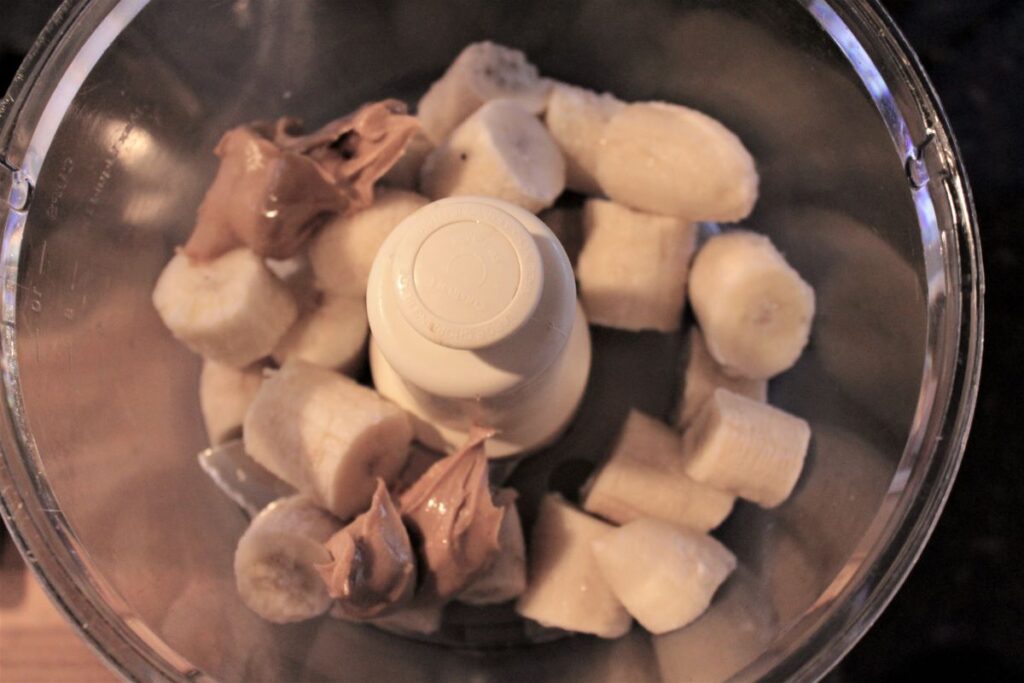 Frozen bananas and peanut butter are placed in the bowl of a food processor.