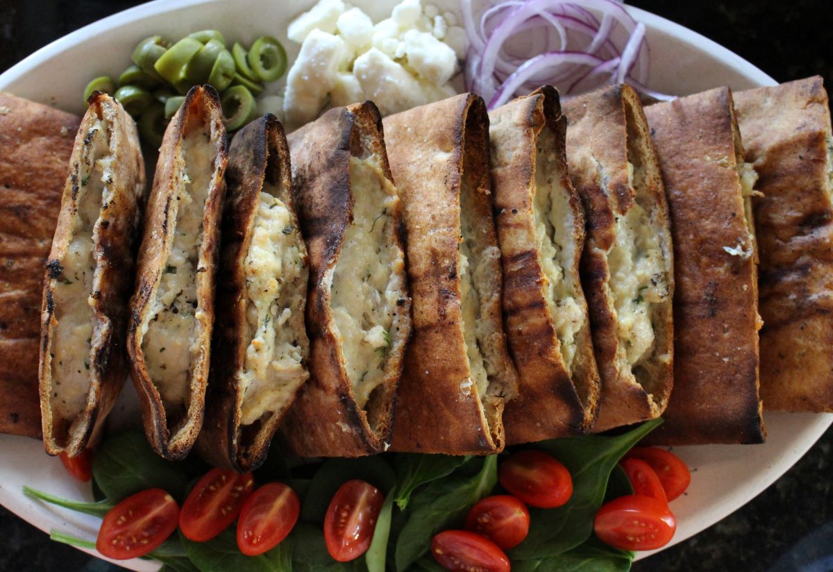 Grilled pita pocket bread that is stuffed with chicken burgers on a platter with condiments: olives, feta, red onion sliced thinly, fresh spinach, and halved cherry tomatoes.