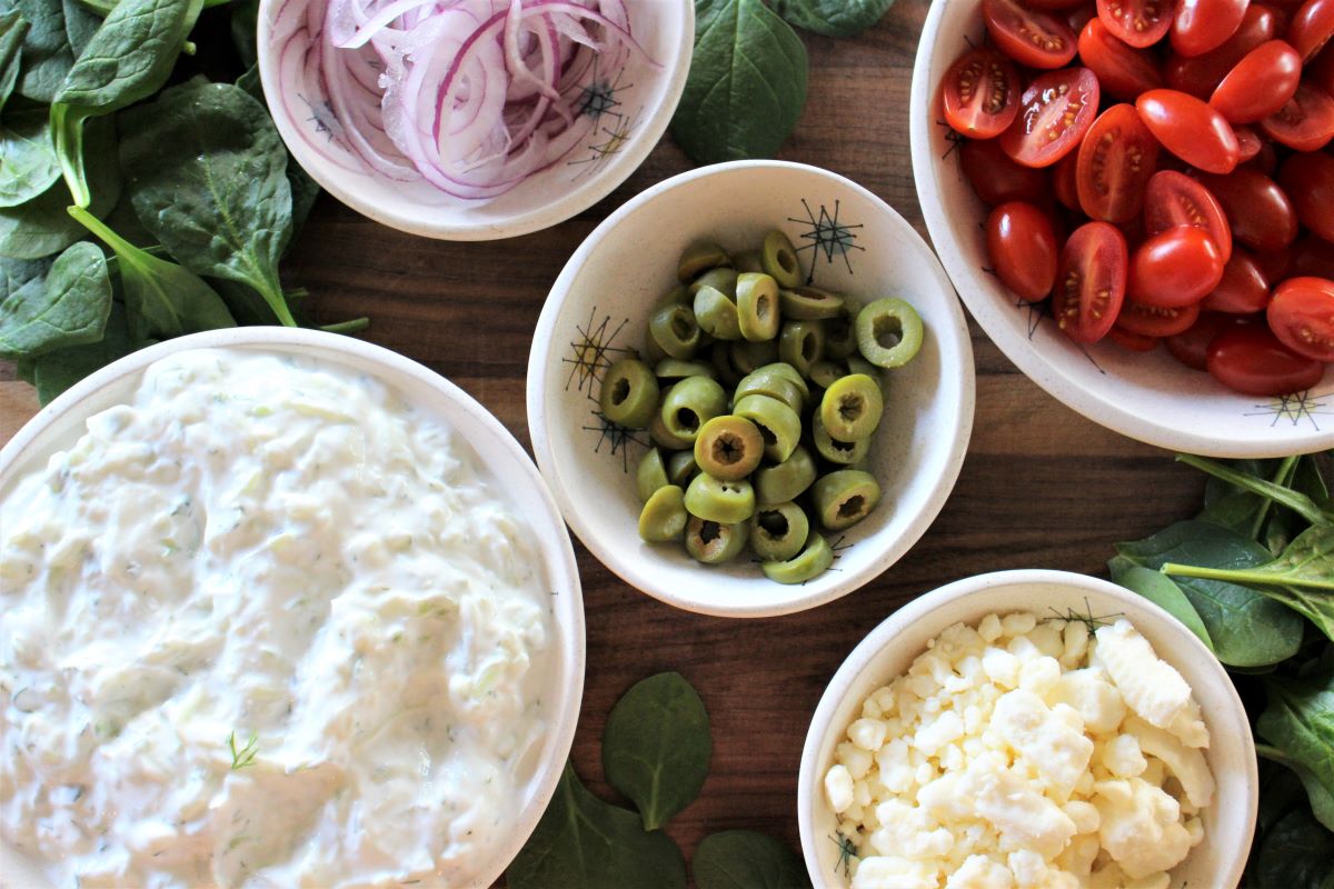 Bowled up condiments for the grilled stuffed pitas. Creamy tzatziki, sliced green olives, crumbled feta cheese, halved cherry tomatoes, thinly sliced red onion, with fresh spinach scattered all around make for a colorful tray of fixings.