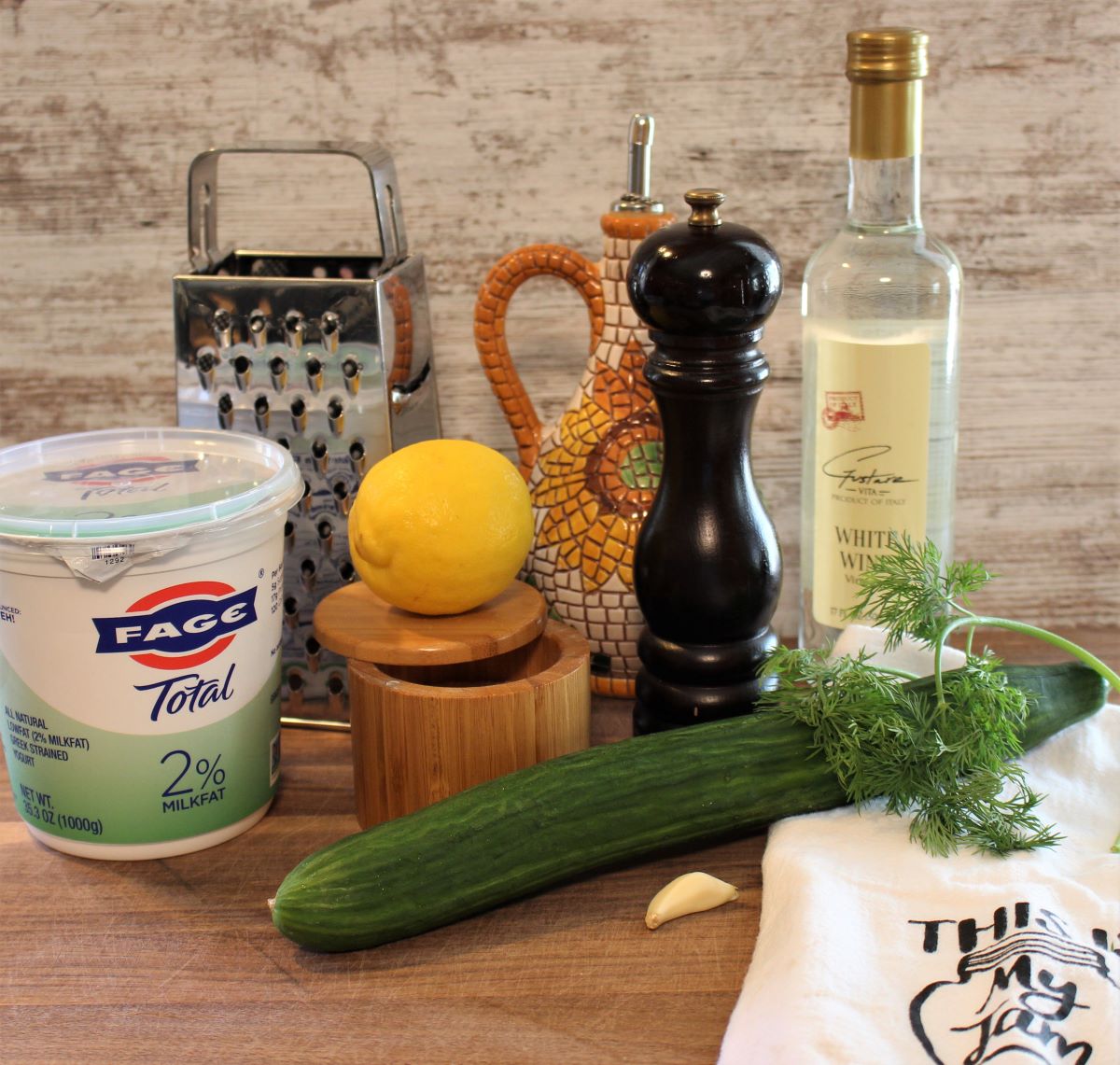 The ingredients needed to make tzatziki for chicken burger pita pockets are Fage Greek Yogurt, lemon juice, cucumbers, white wine vinegar, extra virgin olive oil, garlic, fresh dill, salt and pepper. Supplies needed are a box grater for shredding the cucumber and a tea towel for wringing out the juice of the cucumber.
