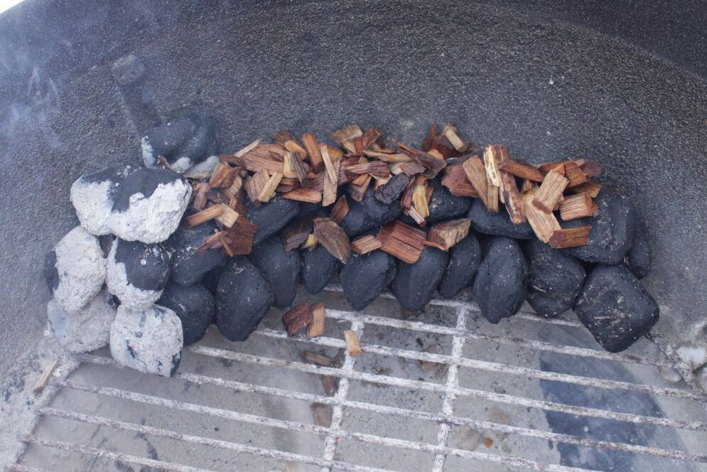 Charcoal is shingled along side of kettle grill. SMoking wood chips are on top of the charcoal. Ashy lit charcoal briquettes are placed on top of the wood chips to create smoke.
