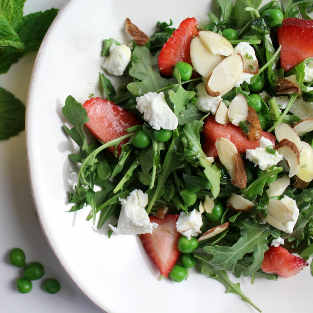 arugula, goat cheese, strawberries, and peas on a white plate with a drizzle of lemony mint dressing sprinkled with sliced almonds