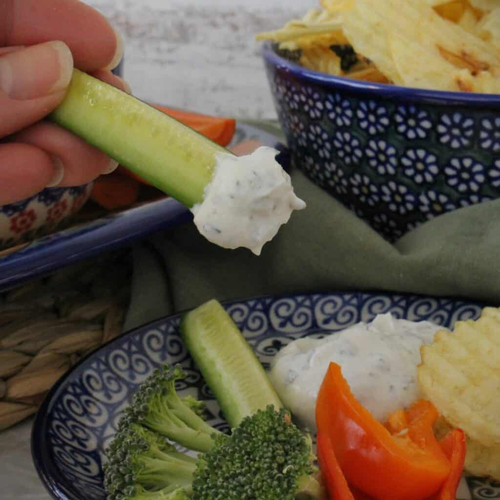 Cut veggies and potato chips on a small plate with a zucchini stick being dipped in healthy ranch dip.