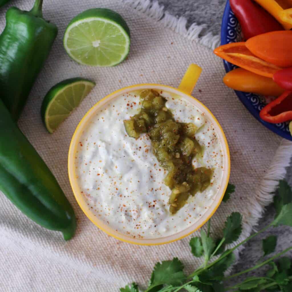healthy queso dip in bowl next to lime wedges, mini bell peppers, green chilies and cilantro.