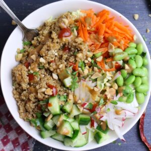 Cooked quinoa, shredded carrots, edamame, diced cucumbers and julienned radishes in a bowl with a drizzle of peanut dressing, a few dabs of sriracha, and sprinkled with chopped peanuts.