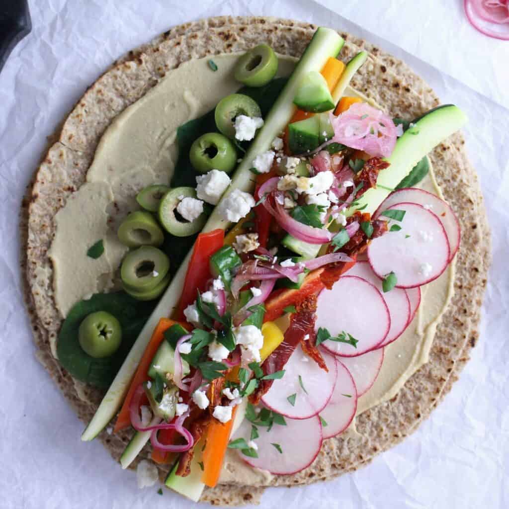 Whole grain wrap with hummus spread on, layered with crunchy veggies, topped with olives, feta, pickled onions, and a sprinkle of fresh parsley.