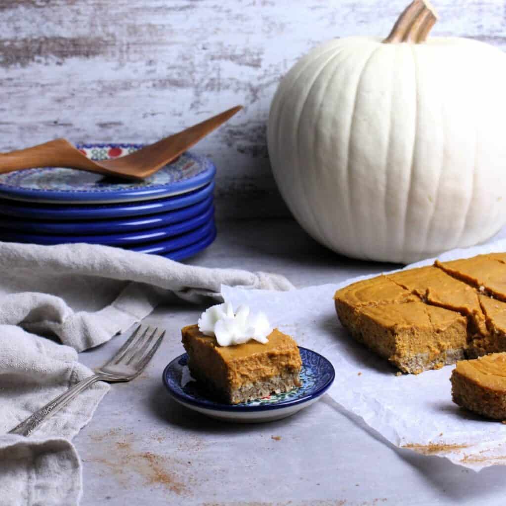 A creamy pumpkin pie bar plated with a white pumpkin in the background, Polish pottery plates, and a wooden server.