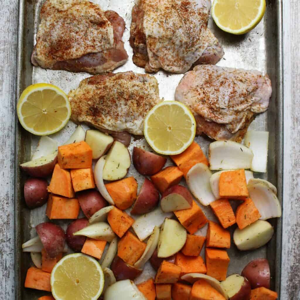 Raw chicken and veggies on a sheet pan with cut lemons ready for roasting.