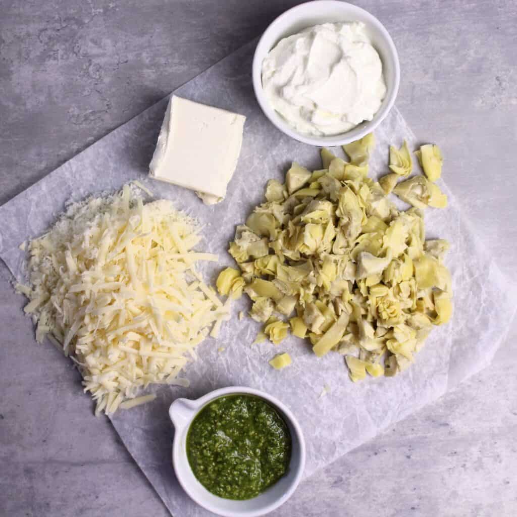 Ingredients needed for Artichoke Pesto Dip are cream cheese, chopped artichokes, Greek yogurt, pesto, and parmesan cheese.