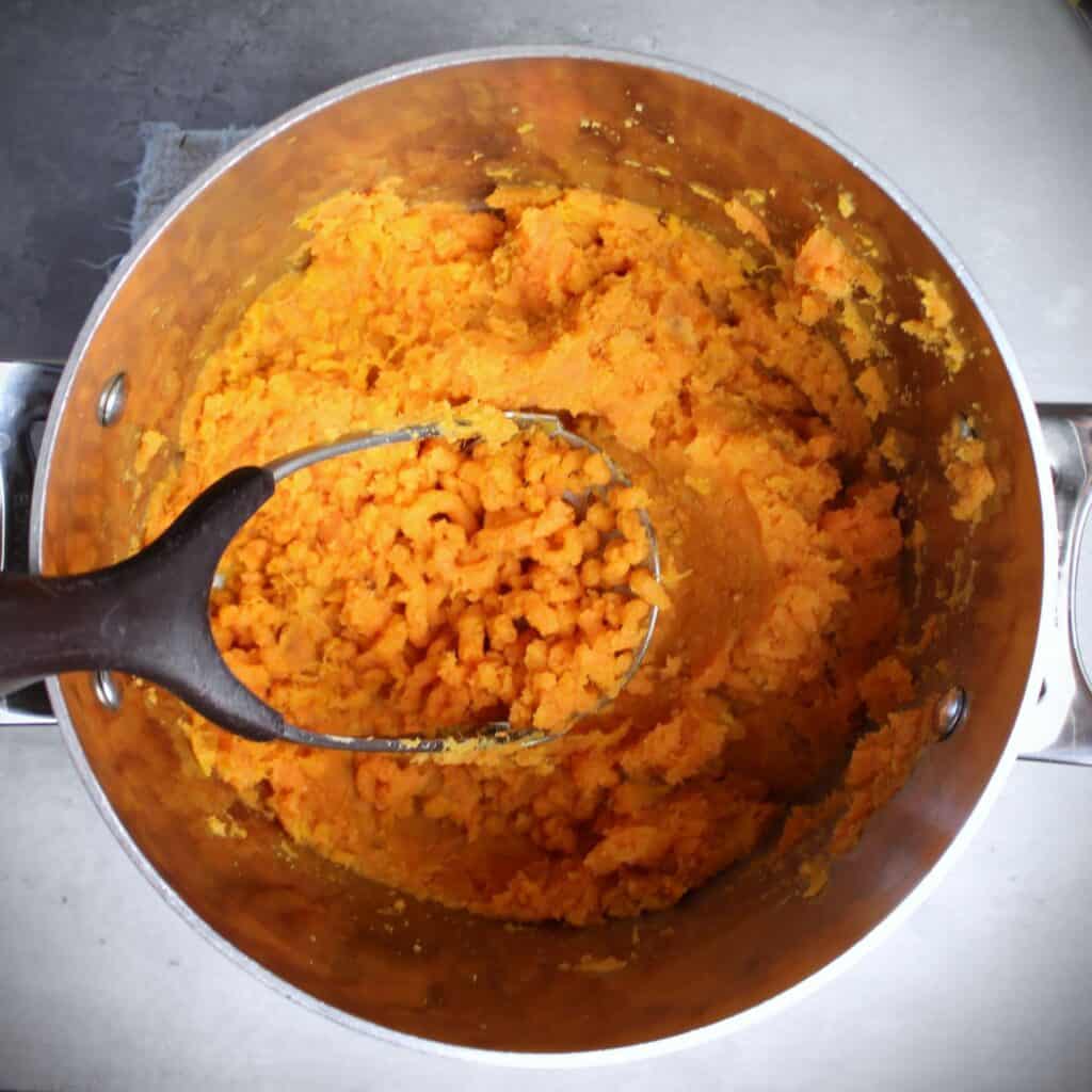 Mashed sweet potatoes in pot with potato masher.