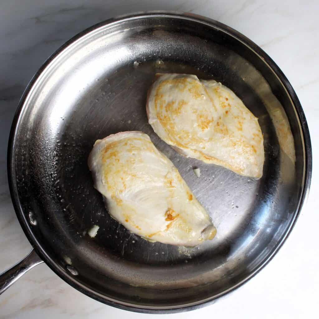 Two chicken breasts being seared in pan.