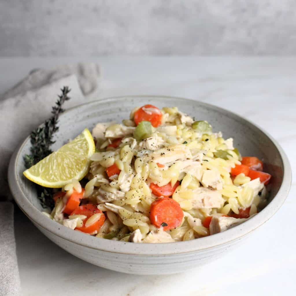 Chicken Orzo Bake in bowl with thyme and lemon wedge .